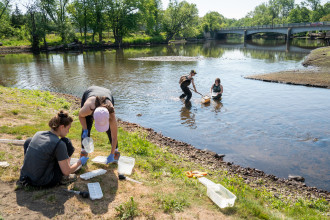 Students record data on the banks of a river while two other students collect samples in the water.