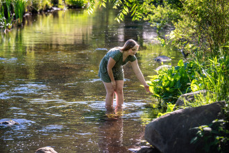 Jossalyn Rogalski wades in a stream and bends down to touch the plants on the water's edge.