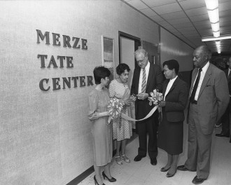 Two people hold a ribbon for Merze Tate to cut in front of the Merze Tate Center.