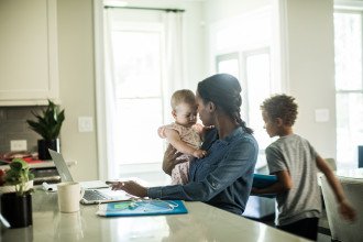 A mom holds a baby while working on her computer on the kitchen counter.