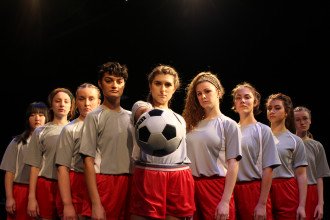A girl's soccer team stands together, with the player in the center holding a soccer ball outstretched.
