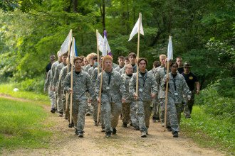 A group of students in Army fatigues marches down a path holding flags.