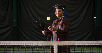 Myint tosses a pickleball in the air getting ready to serve over the net while wearing his graduation cap and gown.