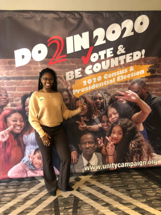 Chiante' Lymon stands in front of a large banner that reads, "Do 2 in 2020, vote and be counted."