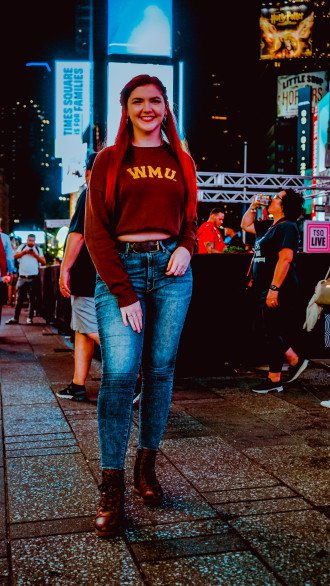 A photo of Grace Stibich standing in Times Square wearing a brown sweater featuring the letters 'WMU' and jeans.