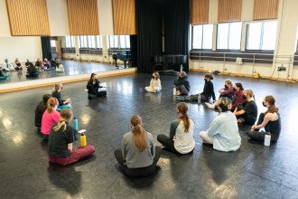 Students sit in a circle on the floor in a dance studio.