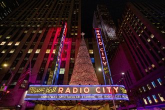 The marquee of Radio City Music Hall lit up at night with a Christmas tree on top.
