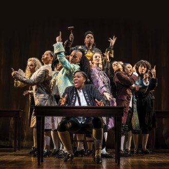 The company of "1776" performs in the production, with Crystal Lucas-Perry seated at a table and the cast behind her.