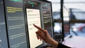 Close-up stock photograph showing a touchscreen monitor being used in an open plan office. A woman’s hand is asking an AI chatbot pre-typed questions &amp; the Artificial Intelligence website is answering.