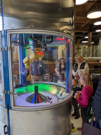 Several people stand around a machine made of glass encasing a robotic arm.