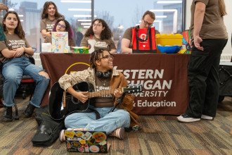 A student sits on the ground playing a guitar.