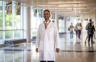 Omer Idris stands in a hallway wearing a white lab coat.