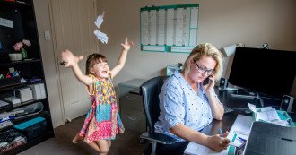 A mom talks on the phone while a child jumps and throws papers in the background.