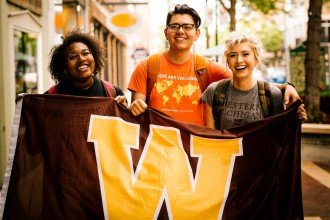 Three students hold a brown and gold "W" flag while standing outside in downtown Kalamazoo.