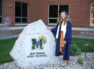 A student in graduation robes standing next to a large rock with the Mattawan High School logo on it.