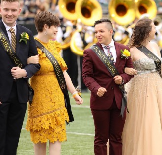 Four WMU students stand on the football field at Waldo Stadium wearing homecoming sashes.