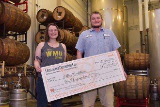 Student Ellie Maddelein with Arcadia brewer Colt Dykstra holding an oversized check for $2,000 and standing in front of brewery barrels.
