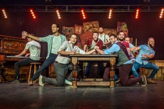 Photo of a group of male performers leaning over a table on a stage with a man playing a piano in the backgroud.