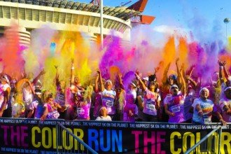 A crowd of runners standing in front of a stadium throwing up colored dust. A banner hangs in front with the words The Color Run, The Happiest 5K on the planet.