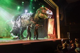 Photo of two actors on a stage with a large Dinosaur prop and people looking on from the floor.