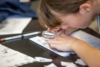 A girl looks at something on a desk closely through a magnifying glass.