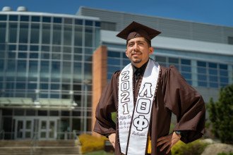 Student Jovanny Ruiz-Alavez stands outside of the Chemistry Building in his graduation cap, gown and sash.