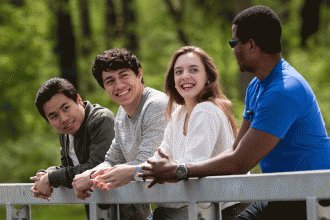 Four smiling students leaning against the railing of a campus bridge.