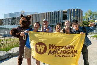 Five students standing in front of the new student center with Buster. They are holding a WMU flag and smiling at the camera.