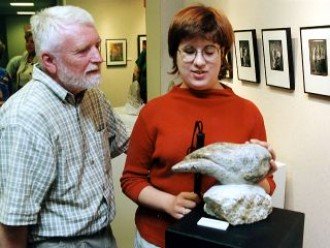 a photo of a professor and a visually impaired student examining what a sculpture. The student has her hand on the sculpture and is feeling the relief carving of the piece of art. It's difficult to tell what the sculpture is from the angle the photo is taken, it may be a small owl, or something entirely abstract.