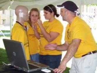 a photo of three students working with a manequin that has a sensor attached to it's head. The students and the manequin are all wearing matching yellow shirts.
