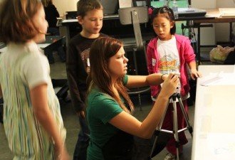 a photo of a student teacher showing elementary students how to use a digital camera in a classroom. The camera is on a tripod which is set at table height to capture an image of a paper that's been placed on the table.