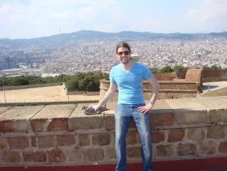 a photo of a graduate student sitting on a wall with a city stretching out behind and below him in the background. 