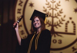 a photo of a student wearing a graduation cap and gown holding her diploma in front of a large wall hanging of the official seal of Western Michigan University
