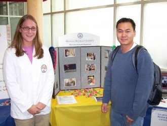 a photo of a medical fair, where two people are standing in front of a fold-out table-top display. There are other tables and displays on each side, but only a very small portion of each is visible. The woman on the left is wearing a white jacket with an emblem on the front pocket and the man on the right is dressed in jeans and a sweatshirt and wearing a backpack.