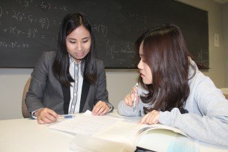 Two asian female students work together, one tutoring the other with the aid of reference books and notes, the board behind them filled with formulas. You too can engage in learning with others at Western Michigan University. 