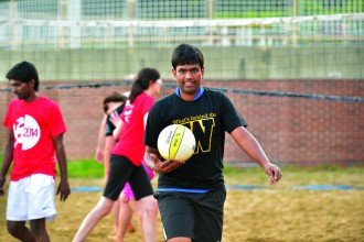 WMU international student in a "W" t-shirt holds a volleyball on the outdoor sand volleyball court