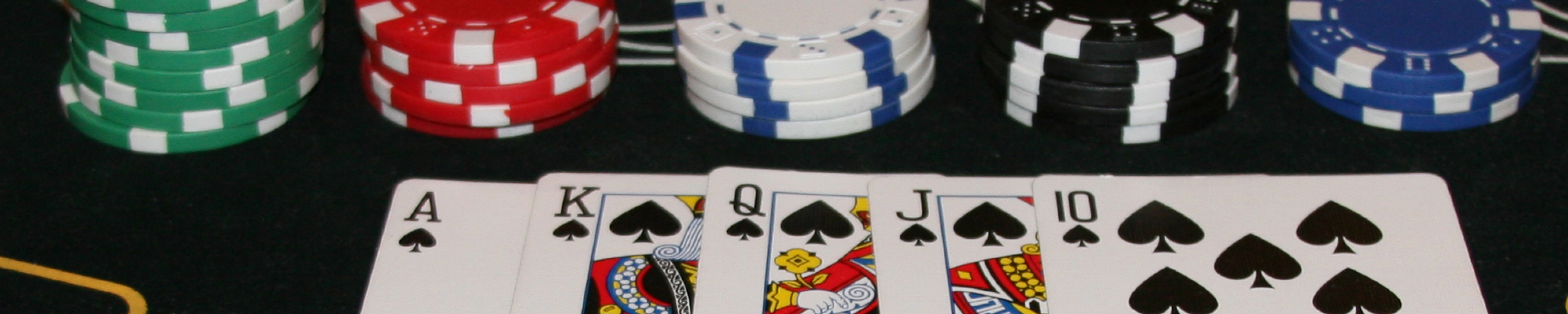 Poker chips in green, red, white and black arranged in a row with a royal flush of cards in Ace, King, Queen, Jack and 10 of spades all laid out in front of chips