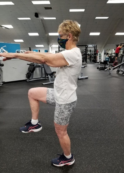 Lady standing and lifting one leg and both arms while in a gym.
