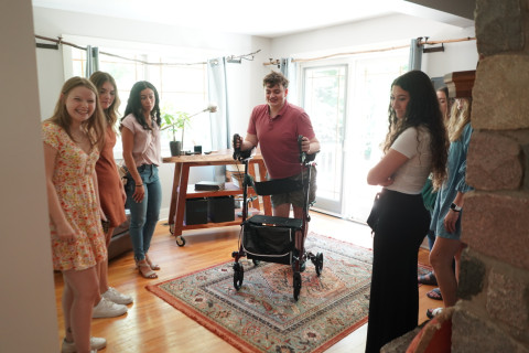 Donovan Lassig uses his walker to navigate a space inside his home while several students look on.