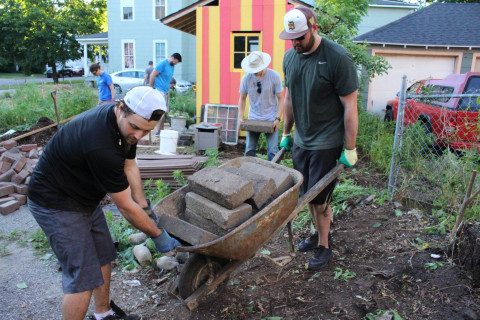 Two young men roll a wheelbarrow full of concrete blocks at a garden construction site.