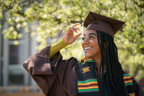A woman in her graduation cap and gown moves her hand to her tassel.