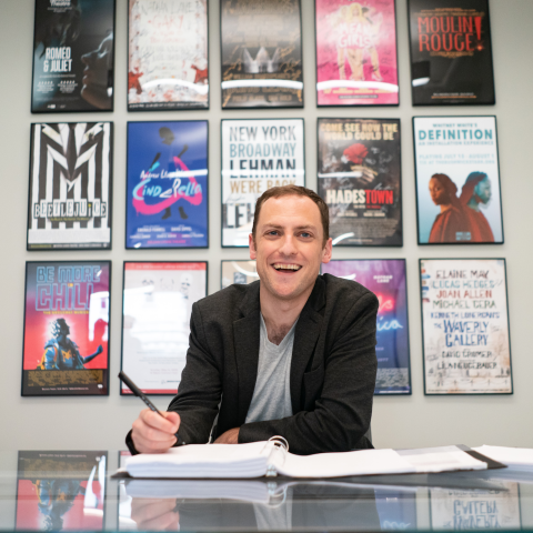 Darren Johnston sits at his desk with a wall of theatre posters in the background.