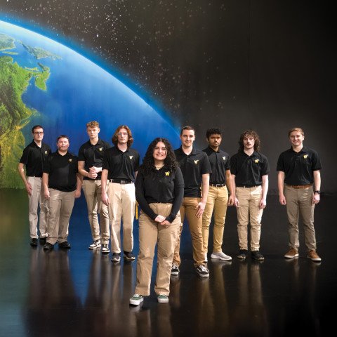 A group of students wearing black shirts and khaki pants stands in front of a wall with art depicting the Earth.