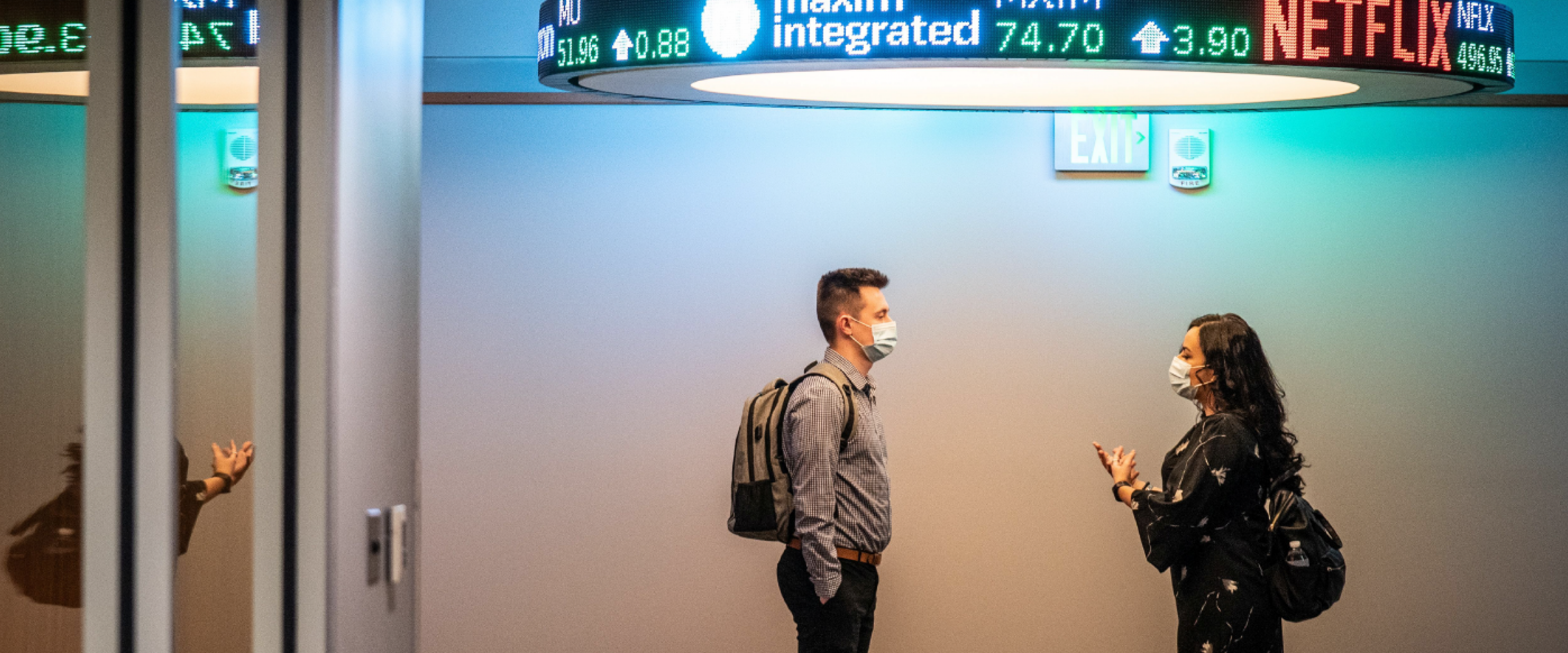 Two masked students standing in hallway under stock ticker