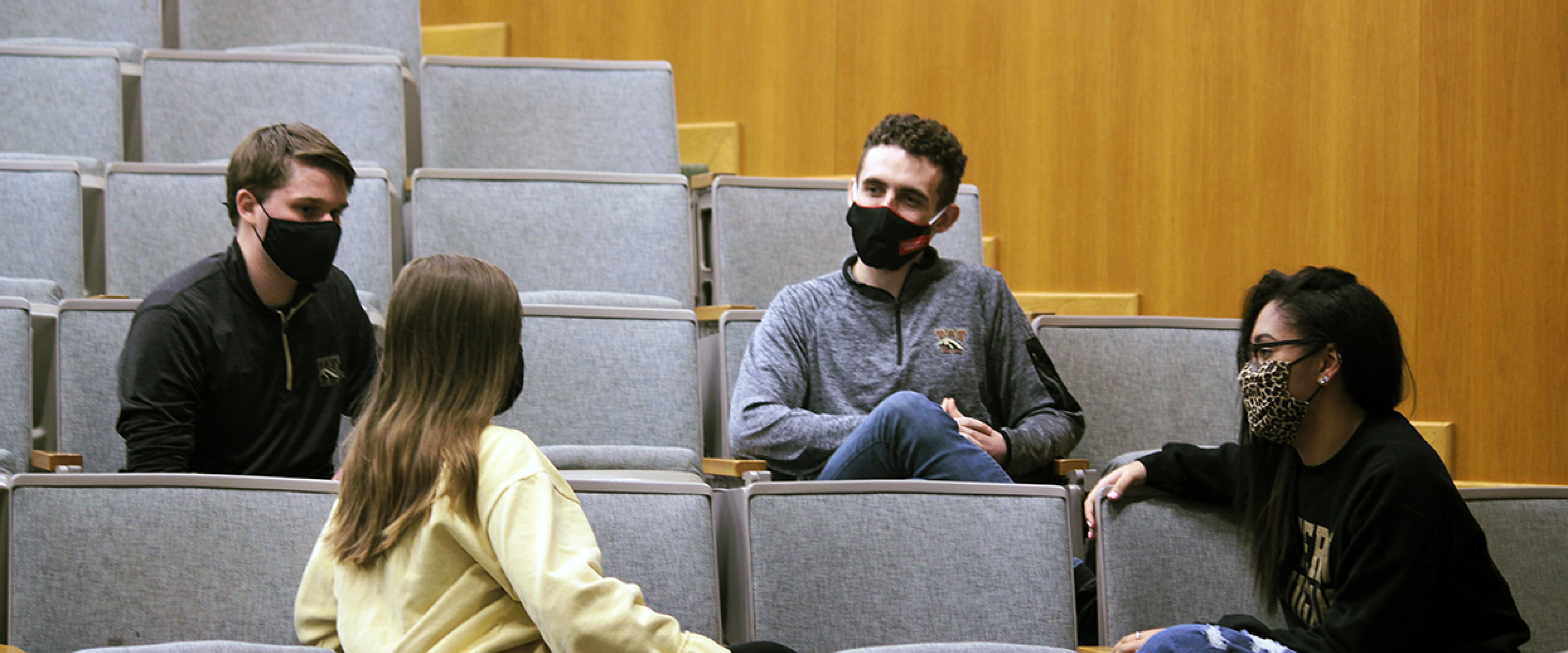 Pictured is a group of masked students, socially distanced, sitting in an auditorium