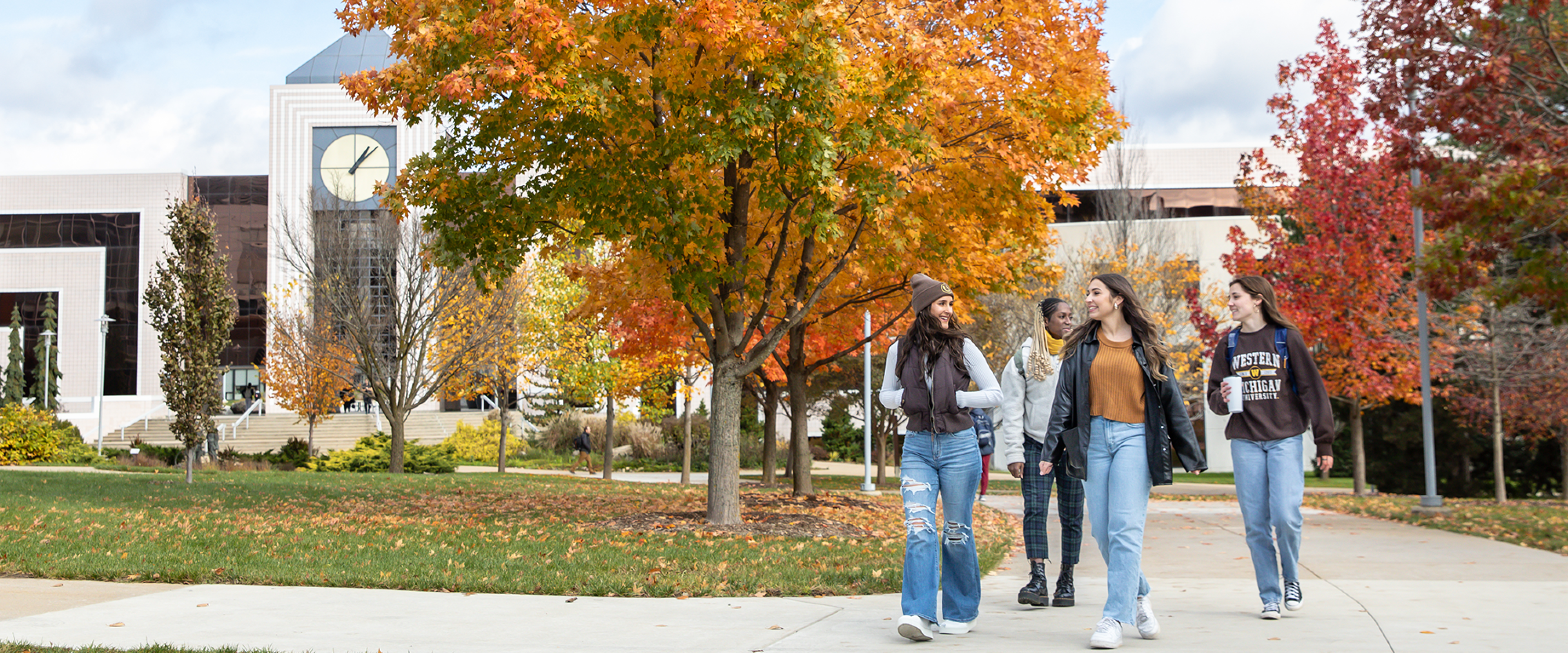 Group of WMU students walking on campus sidewalks in front of popular building and fall colored trees.