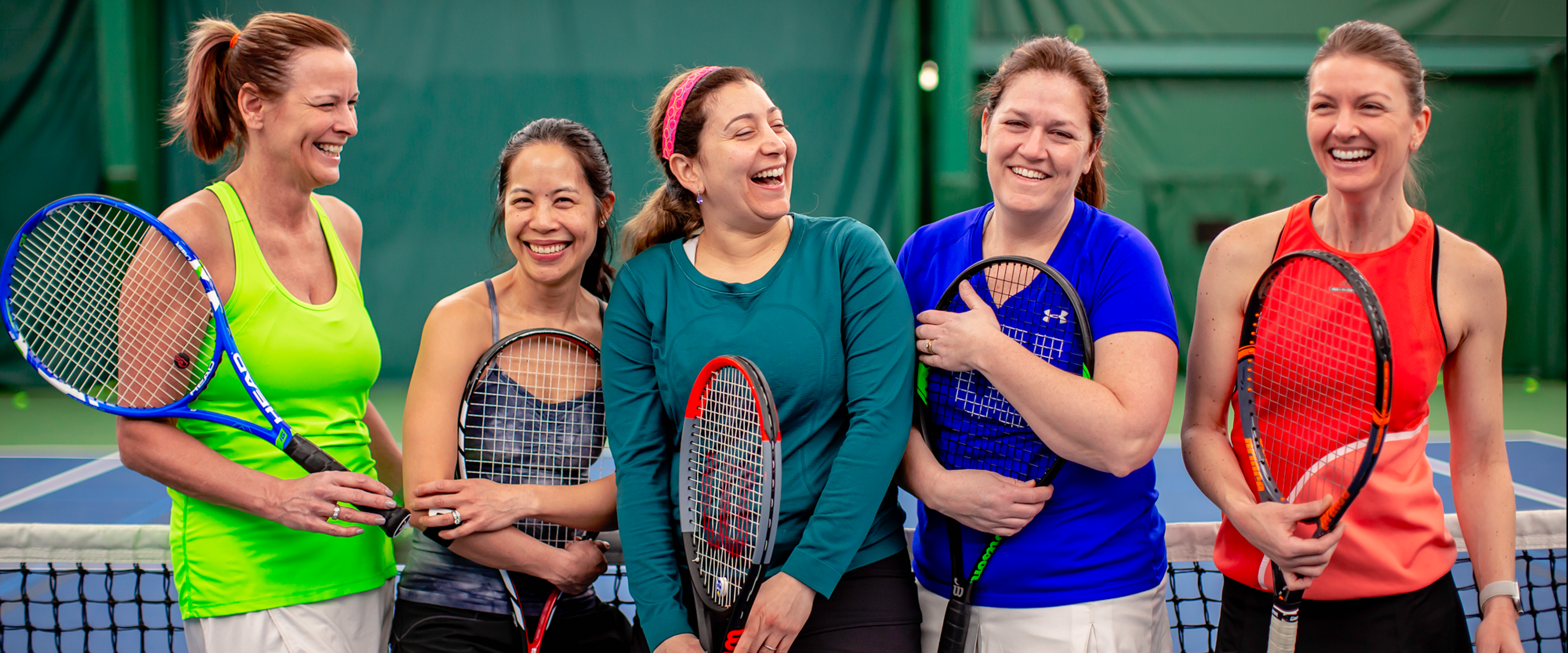 A bunch of ladies at the net holding their tennis rackets and laughing.