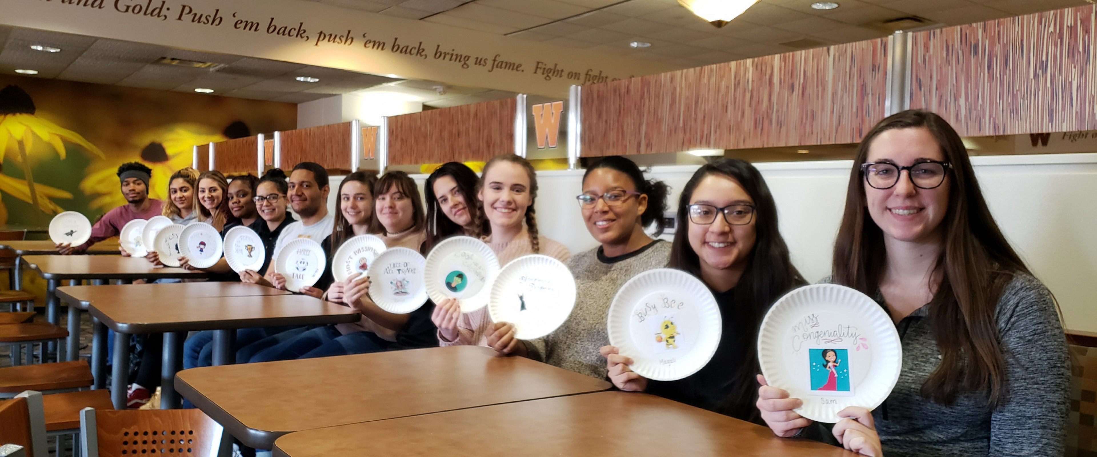 A row of 13 students sitting down, holding up their paper plate awards and smiling at the camera.