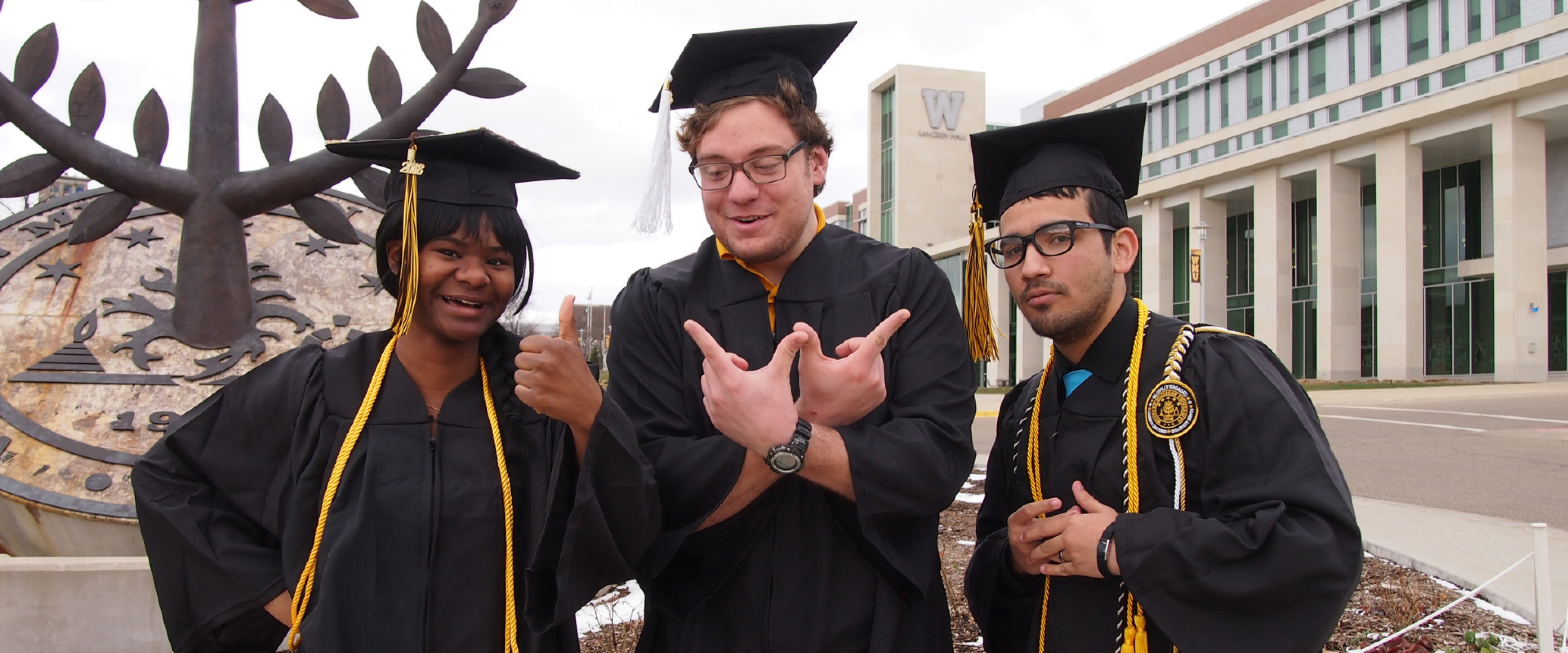 Three students standing in front of the WMU seal statue in their caps and gowns. One student is forming the letter "W" with four fingers.