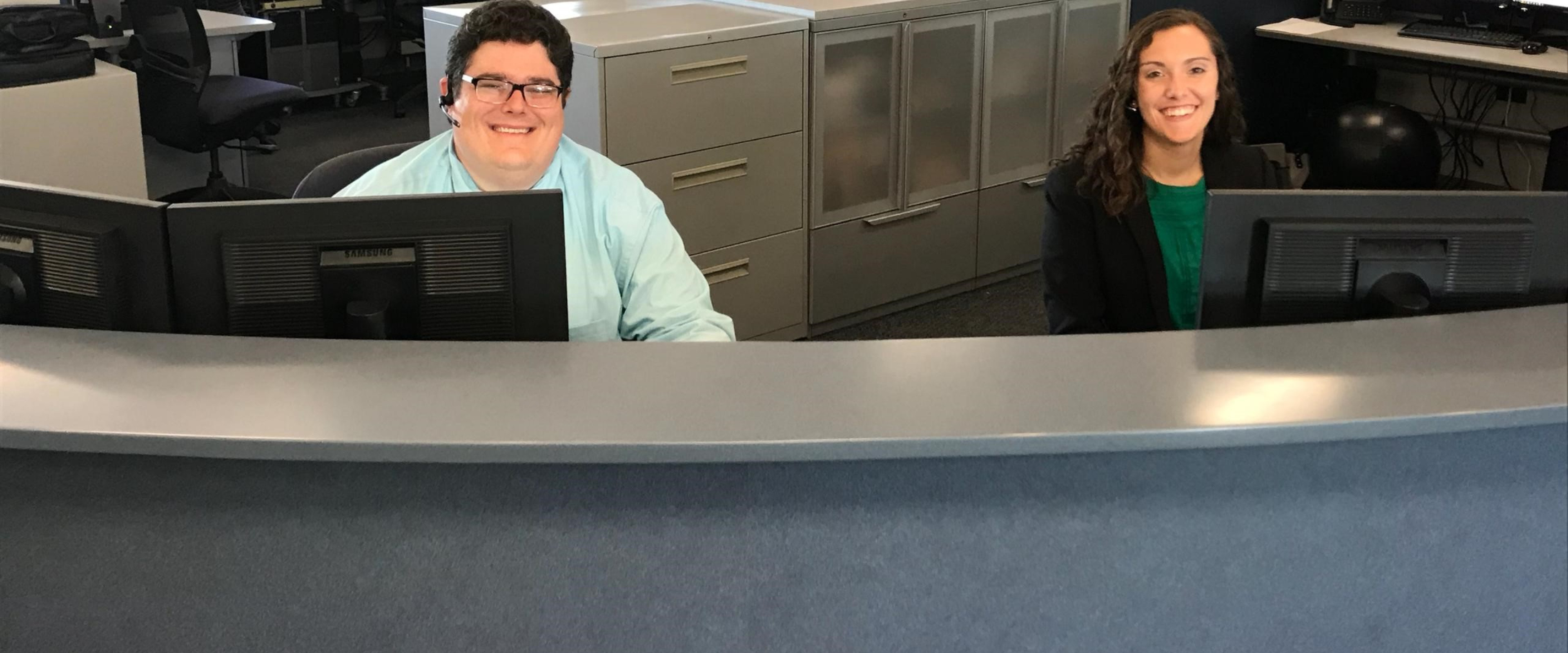 Two employees sit at a desk behind computers with cabinets and more computers behind them